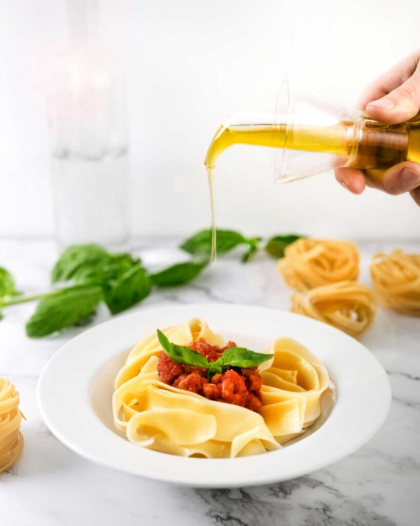 Ein Schuss Olivenöl wird über einen Teller frischer Tagliatelle mit Tomatensugo und Basilikum gegossen – im Hintergrund frische Basilikumblätter und ungekochte Pasta-Nester.
