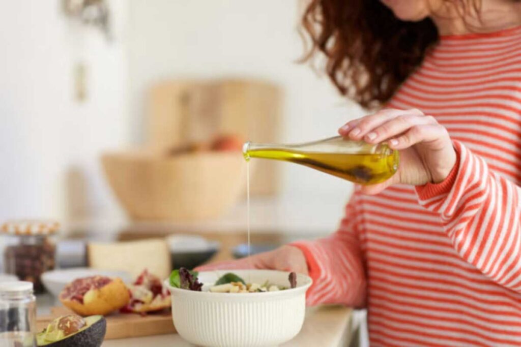 Frau in rot-weiß gestreiftem Pullover gießt natives Olivenöl extra über einen frischen Salat in einer hellen Küche – gesunde mediterrane Küche zu Hause.