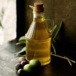 Flasche mit Olio Extra Vergine, daneben frische grüne und dunkle Oliven auf einem Tisch im natürlichen Licht.