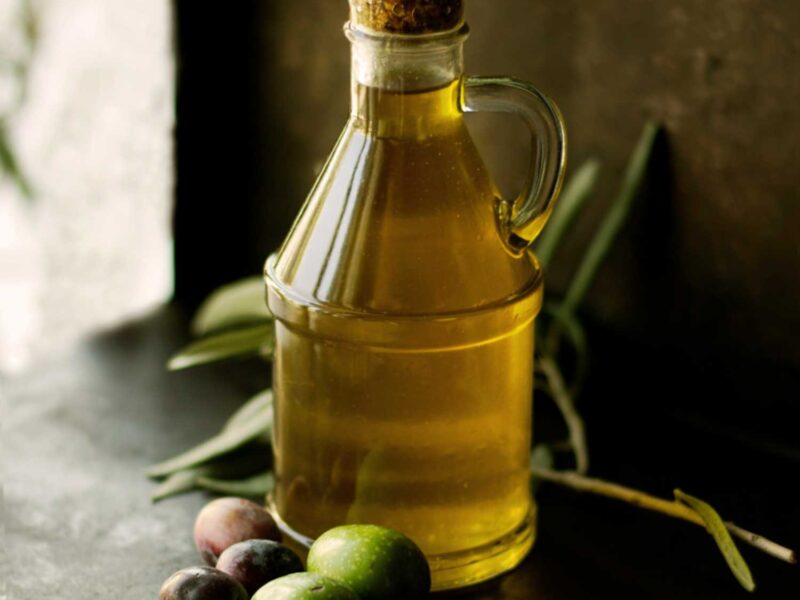 Flasche mit Olio Extra Vergine, daneben frische grüne und dunkle Oliven auf einem Tisch im natürlichen Licht.