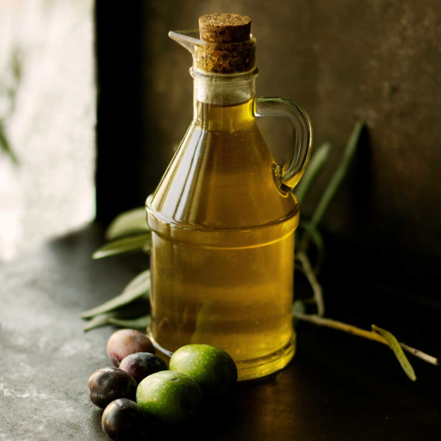 Flasche mit Olio Extra Vergine, daneben frische grüne und dunkle Oliven auf einem Tisch im natürlichen Licht.