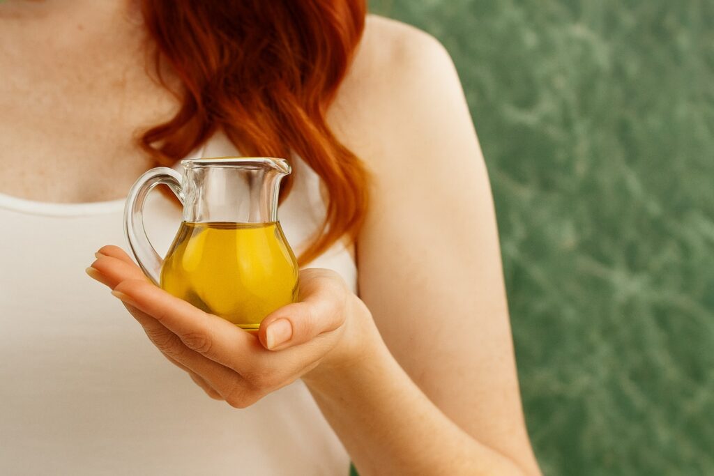 Frau mit roten Haaren hält eine kleine Glaskanne mit goldenem Olivenöl in der Hand – grüner Hintergrund.