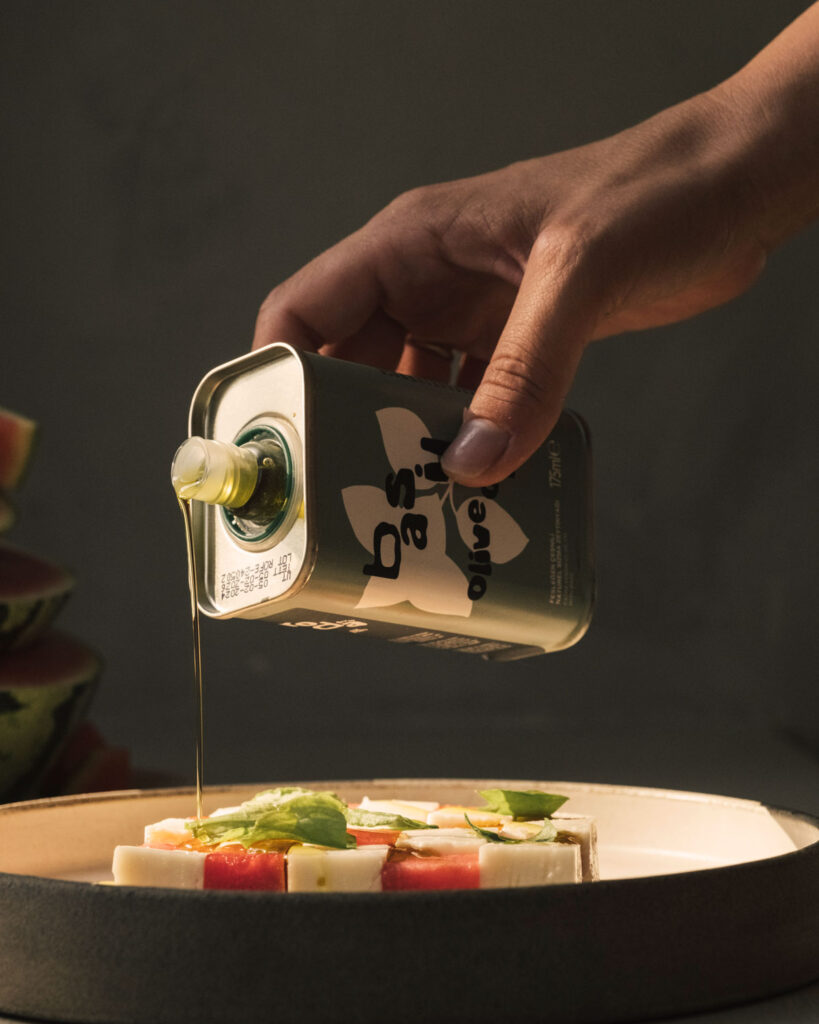 Olivenöl wird aus einer Designer-Metallflasche auf eine Caprese mit Wassermelone und Basilikum geträufelt.