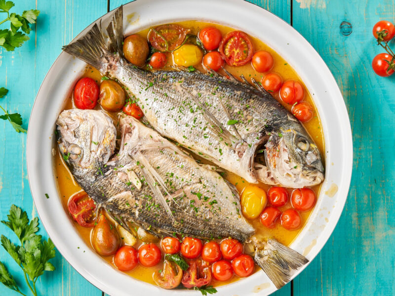 Zwei ganze Doraden in aromatischem Sud mit bunten Tomaten auf hellblauem Holz – mediterrane Authentizität pur aus der italienischen Küche.