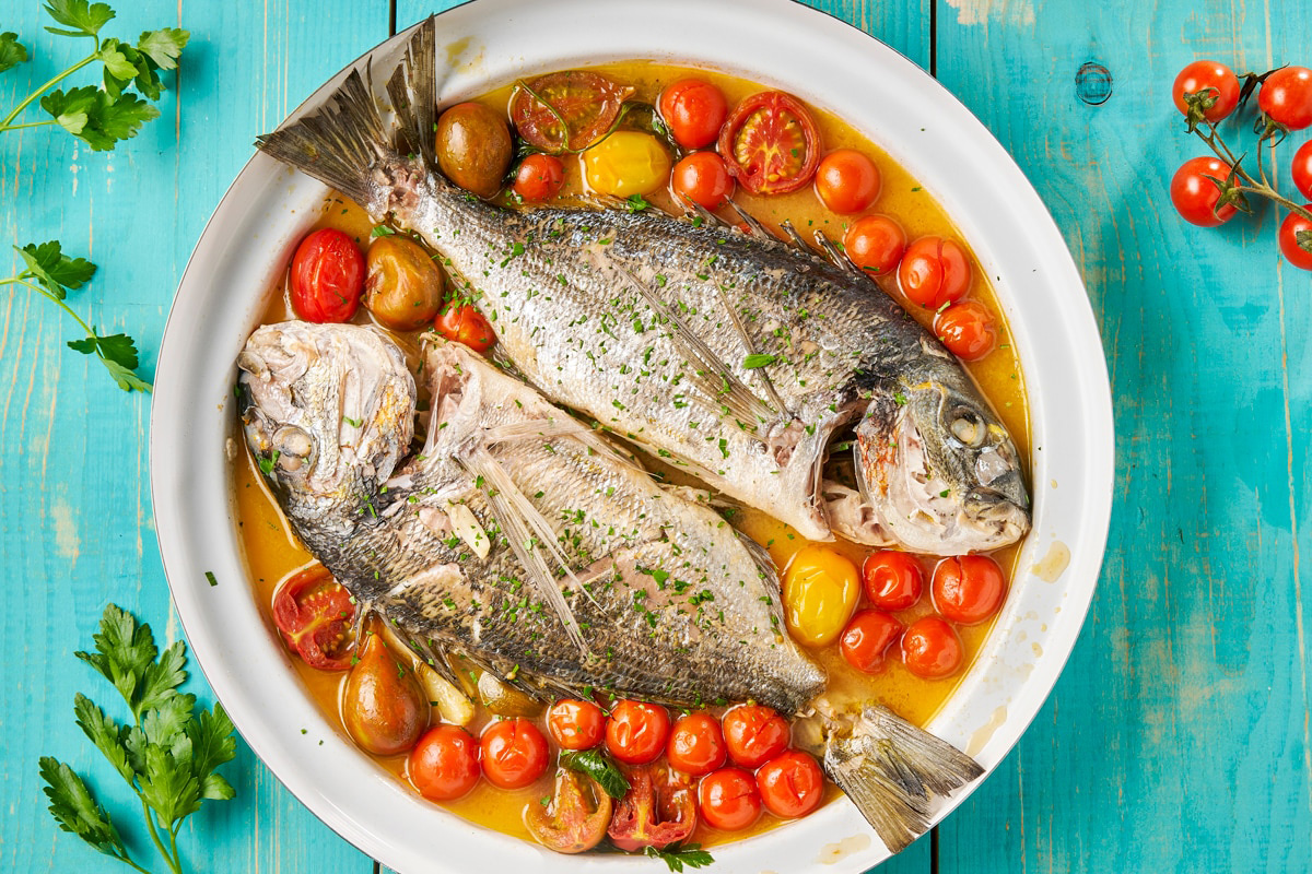 Zwei ganze Doraden in aromatischem Sud mit bunten Tomaten auf hellblauem Holz – mediterrane Authentizität pur aus der italienischen Küche.