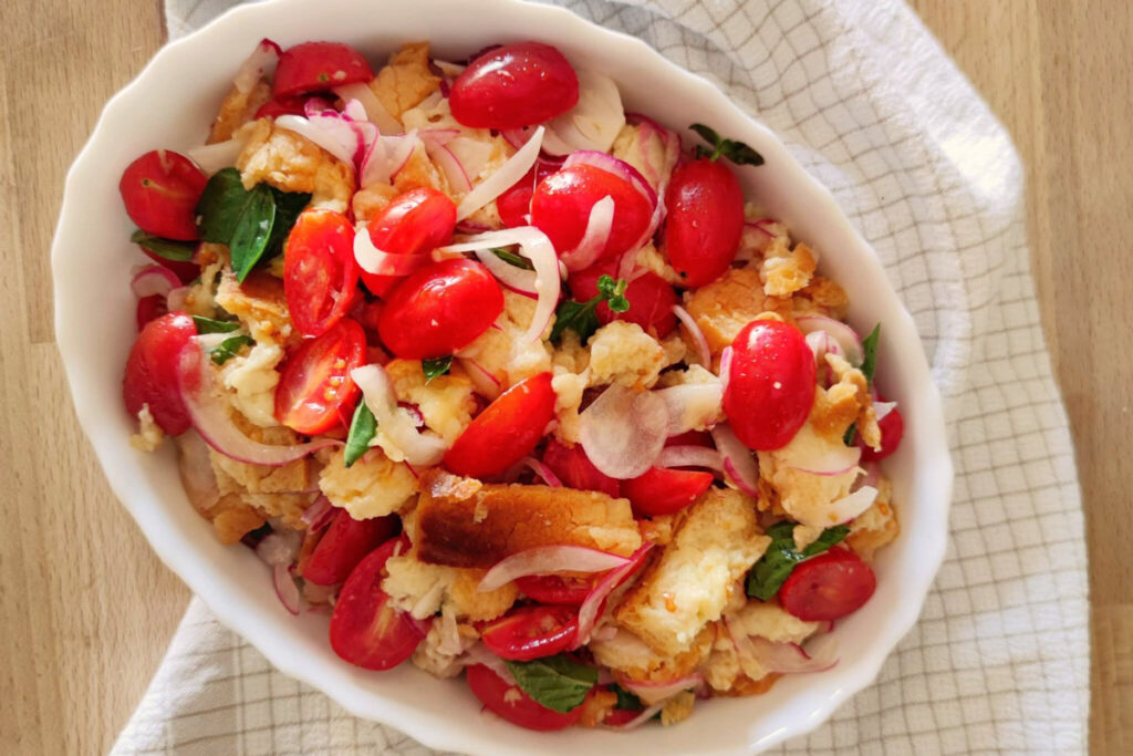 Nahaufnahme einer traditionellen Panzanella mit Kirschtomaten, roten Zwiebeln, Basilikum und eingeweichtem Brot in einer weißen Schüssel.