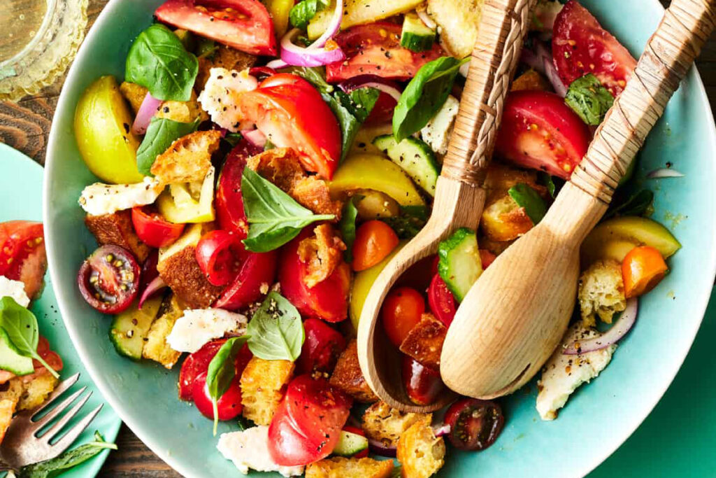 Bunte Sommer-Panzanella mit verschiedenen Tomatensorten, Basilikum, Brotstücken und roten Zwiebeln – angerichtet in einer türkisen Schüssel mit Holzbesteck.