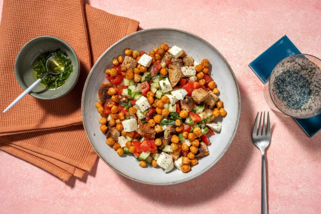 Moderne Interpretation der Panzanella mit gerösteten Kichererbsen, Feta, Gurken, Tomaten und geröstetem Brot auf rosa Hintergrund.