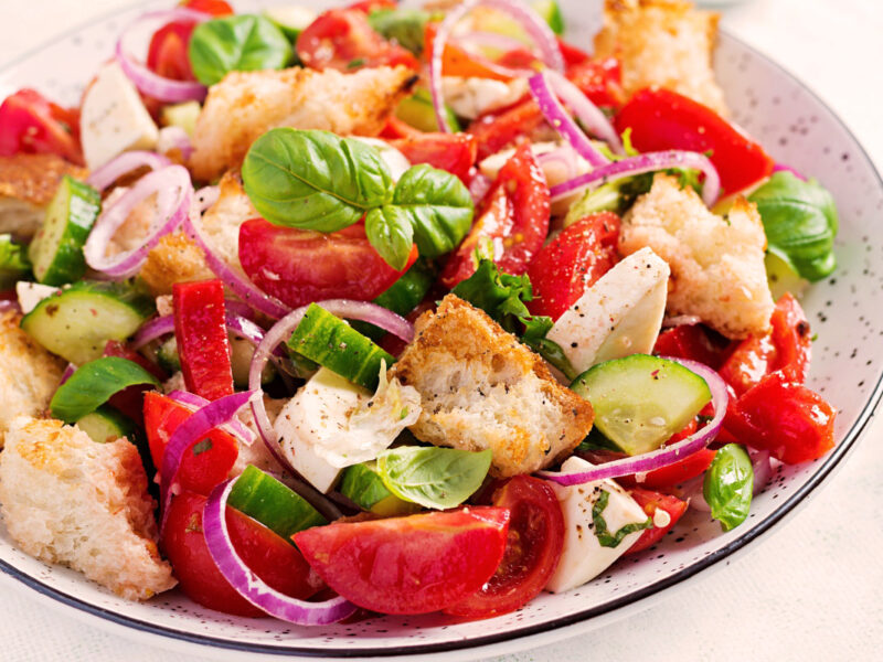 Frische Panzanella mit Tomaten, Gurken, roten Zwiebeln, Mozzarella, Basilikum und knusprigem Brot – serviert auf einem weißen Teller mit schwarzem Rand.