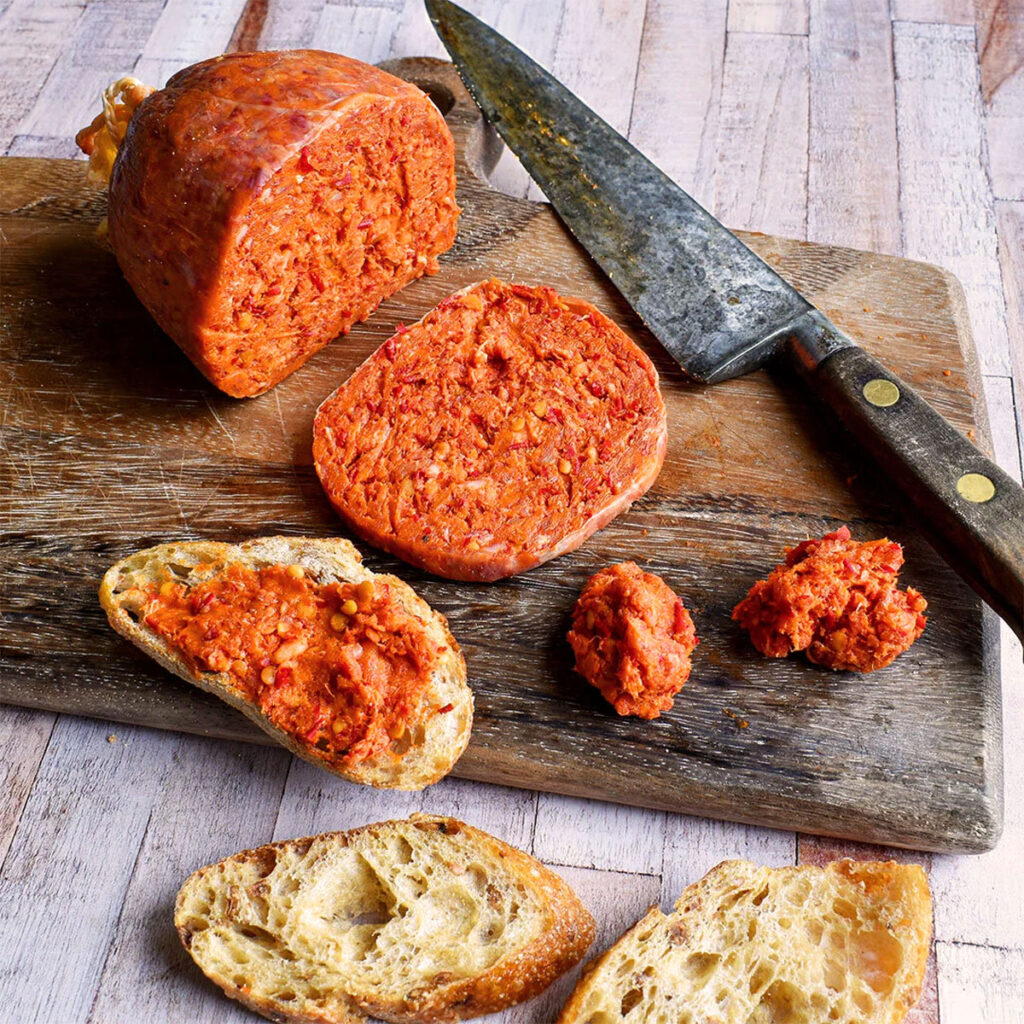 Hausgemachte ’Nduja aus Kalabrien, aufgeschnitten auf einem rustikalen Holzbrett, daneben ein scharfes Küchenmesser und frisches Brot mit Aufstrich.