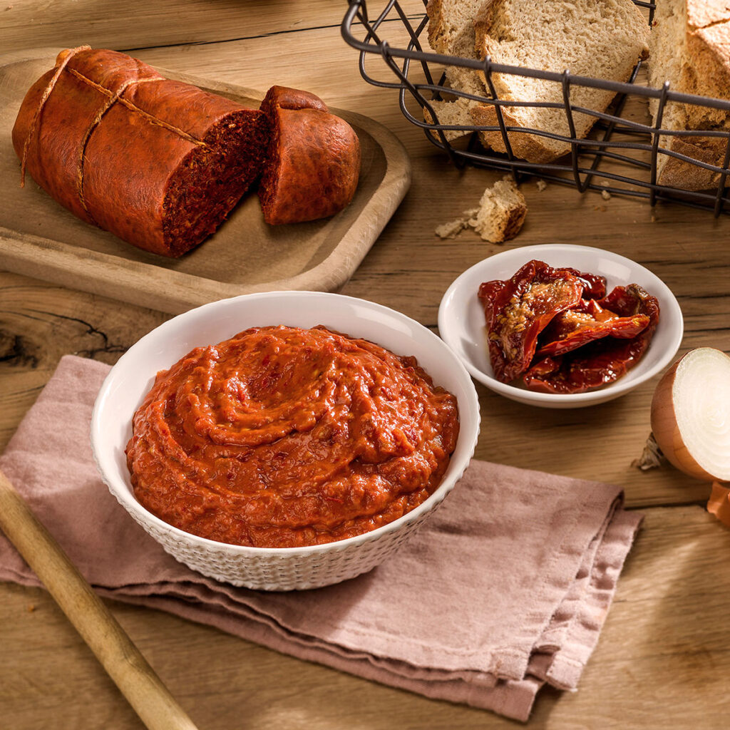 Cremige ’Nduja als würziger Brotaufstrich in einer weißen Schale, serviert mit getrockneten Tomaten, Brot und roher Zwiebel – typisch kalabrisch.