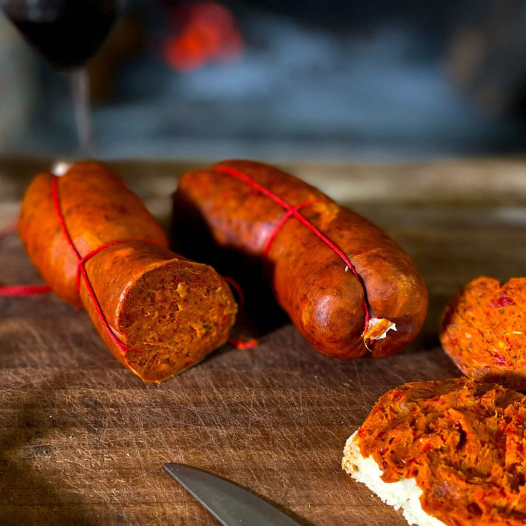 Traditionelle kalabrische ’Nduja-Wurst aufgeschnitten auf einem Holzbrett, im Hintergrund ein Kaminfeuer – rustikale Atmosphäre aus Süditalien.