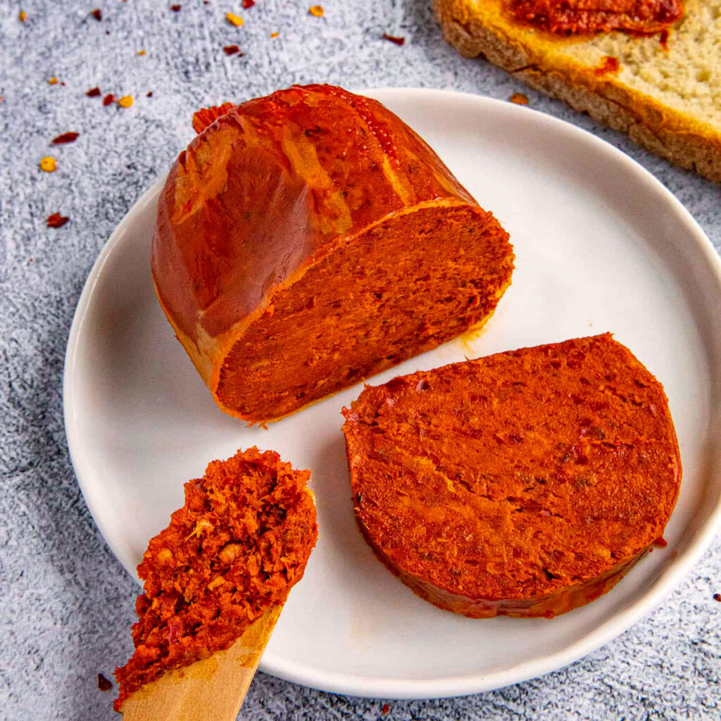 ’Nduja-Wurst aufgeschnitten auf einem weißen Teller, daneben eine Holzlöffelportion zum Aufstreichen – scharf, weich und aromatisch.