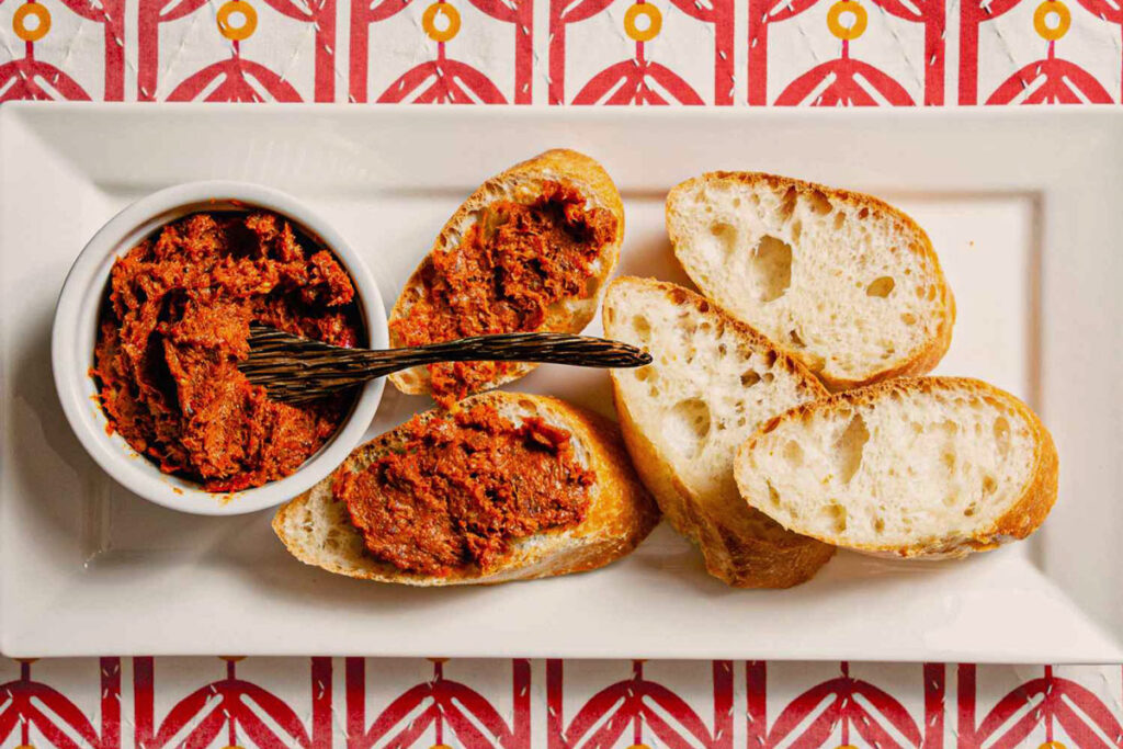 Crostini mit würzigem ’Nduja-Aufstrich auf einem weißen Teller, daneben frisches Baguette und ein kleiner Servierteller – ideal als Antipasto.