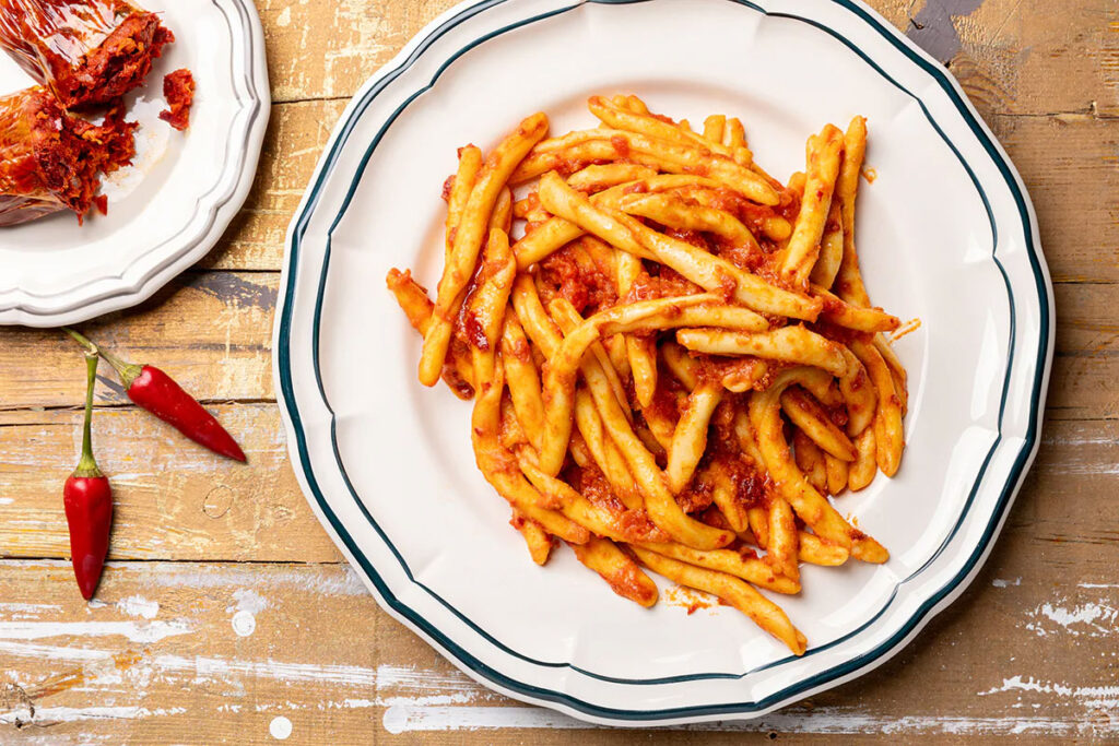 Teller mit kalabrischen Fileja-Nudeln, serviert mit einer kräftigen ’Nduja-Tomatensauce, daneben Chilischoten und aufgeschnittene Wurst.