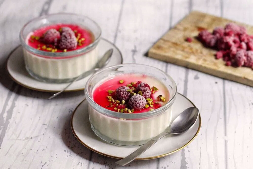 Zwei Gläser Panna Cotta mit Himbeersauce, gefrorenen Himbeeren und gehackten Pistazien, serviert auf weißen Untertassen mit Löffeln.
