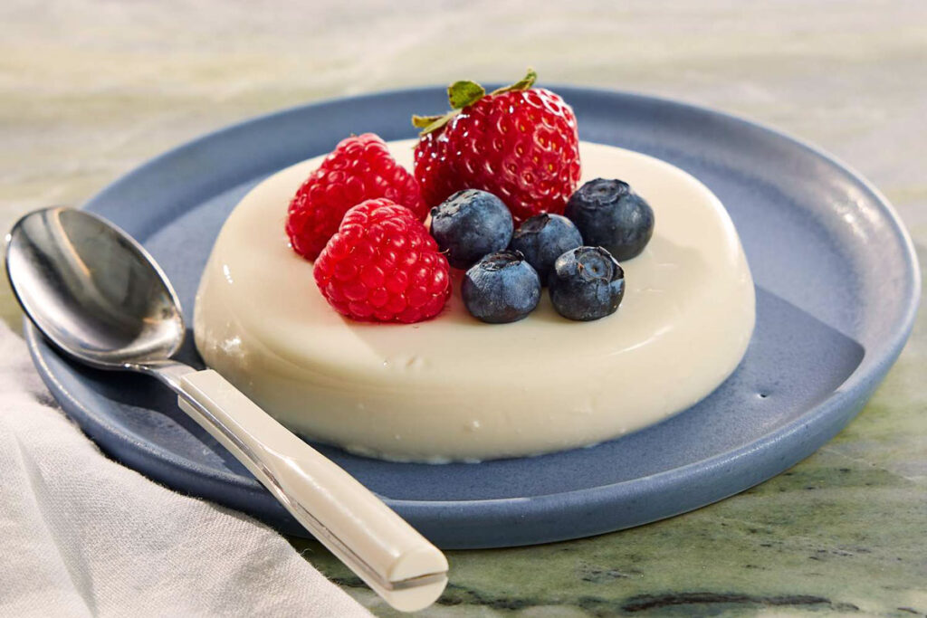 Klassische Panna Cotta auf einem blauen Teller, dekoriert mit frischen Erdbeeren, Himbeeren und Heidelbeeren.