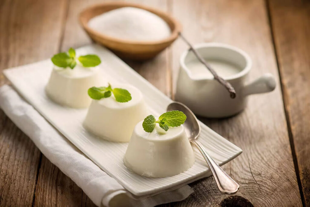 Drei klassische Panna Cotta Portionen auf einem weißen Porzellanteller, verziert mit frischen Minzblättern, daneben eine Kanne Sahne und eine Schote Vanille – rustikal serviert auf Holztisch.