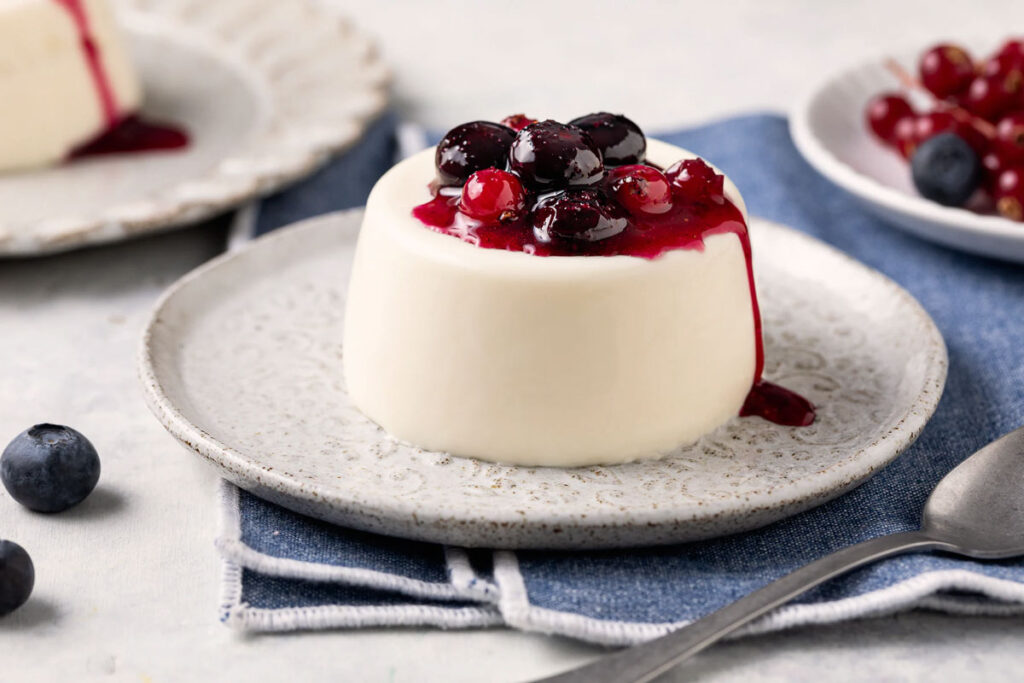 Klassische Panna Cotta auf einem Teller, serviert mit einer intensiven roten Beerensauce und frischen schwarzen Johannisbeeren.