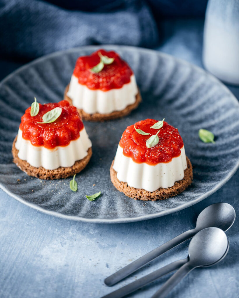 Panna Cotta auf knusprigem Keksboden mit einer glänzenden Erdbeercreme-Gelee-Schicht, dekorativ serviert auf einem dunklen Teller – eine fruchtige und elegante Dessertkreation.