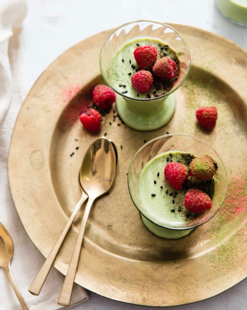 Matcha-Panna-Cotta mit frischen Himbeeren und schwarzem Sesam, elegant serviert auf einem goldenen Tablett mit goldenen Löffeln.