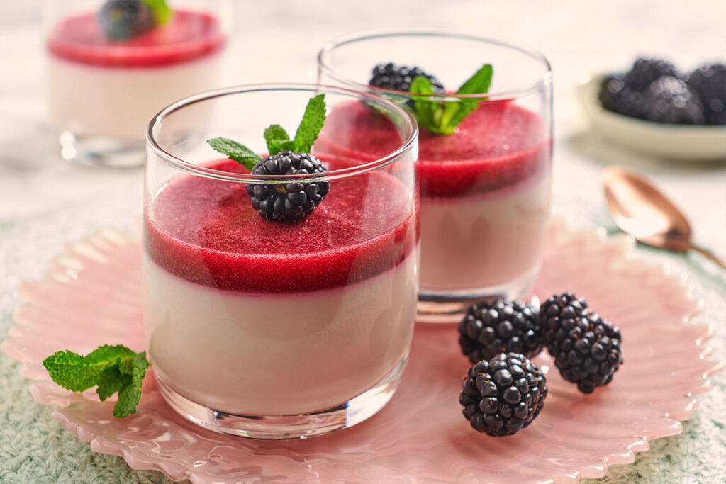 Panna Cotta im Glas mit Brombeerspiegel, garniert mit frischer Minze und Brombeeren, serviert auf einem rosa Teller.