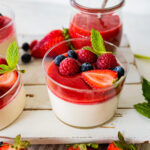 Glasdesserts mit Panna Cotta und leuchtend rotem Beerenspiegel, garniert mit frischen Himbeeren, Heidelbeeren, Erdbeeren und Minze – sommerlich-fruchtige Variante.