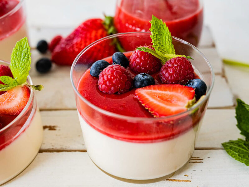 Glasdesserts mit Panna Cotta und leuchtend rotem Beerenspiegel, garniert mit frischen Himbeeren, Heidelbeeren, Erdbeeren und Minze – sommerlich-fruchtige Variante.
