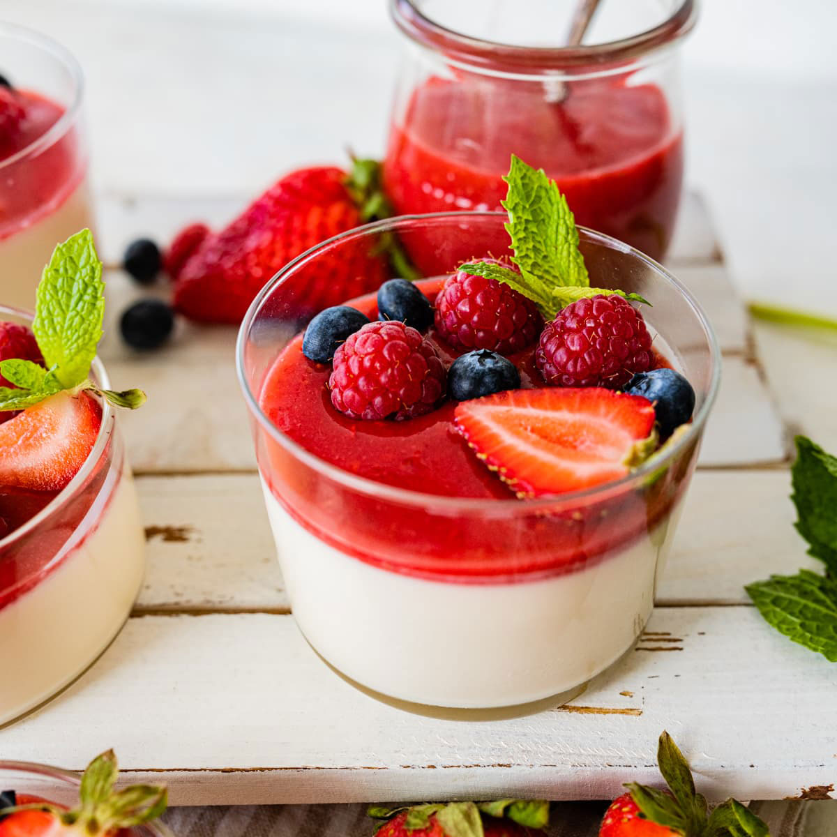 Glasdesserts mit Panna Cotta und leuchtend rotem Beerenspiegel, garniert mit frischen Himbeeren, Heidelbeeren, Erdbeeren und Minze – sommerlich-fruchtige Variante.