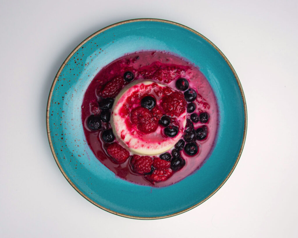 Panna Cotta mit Waldbeeren-Sauce auf einem türkisfarbenen Teller – eine elegante italienische Nachspeise mit frischer Beerennote.