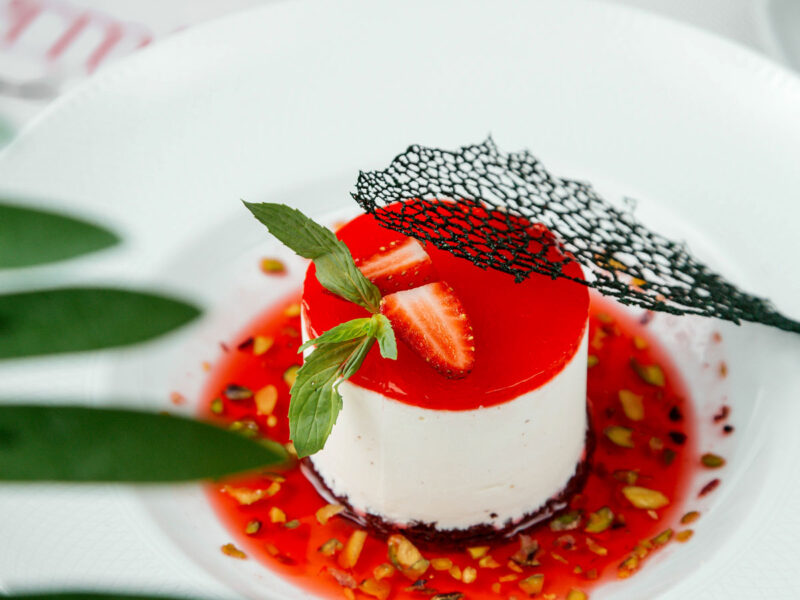 Fein angerichtete Panna Cotta mit Erdbeerspiegel, frischer Minze, Pistazienstücken und schwarzem Netzdekor auf weißem Porzellanteller.