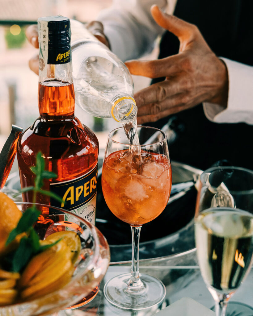 Ein Barkeeper gießt sprudelndes Wasser in ein Glas mit Eis und Aperol auf einem eleganten Tisch – die klassische Zubereitung eines Spritz im italienischen Stil.