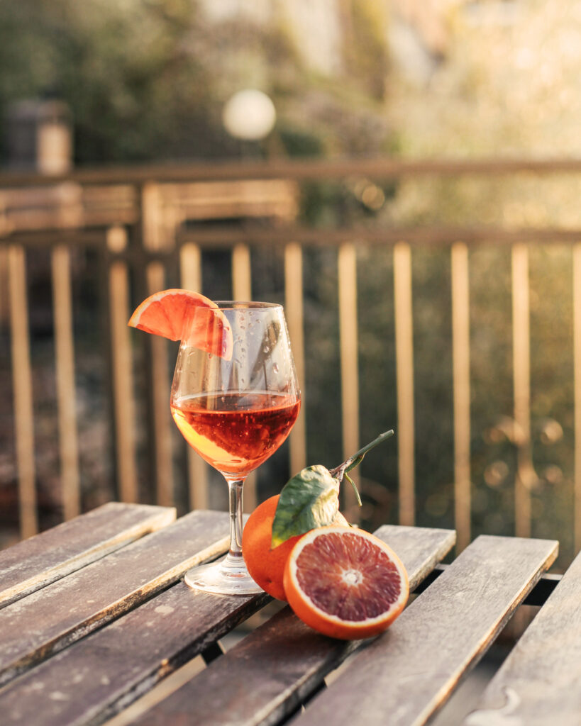Ein Glas Spritz mit Blutorangenscheibe steht auf einem rustikalen Holztisch, begleitet von frischen Blutorangen und mediterranem Abendlicht.