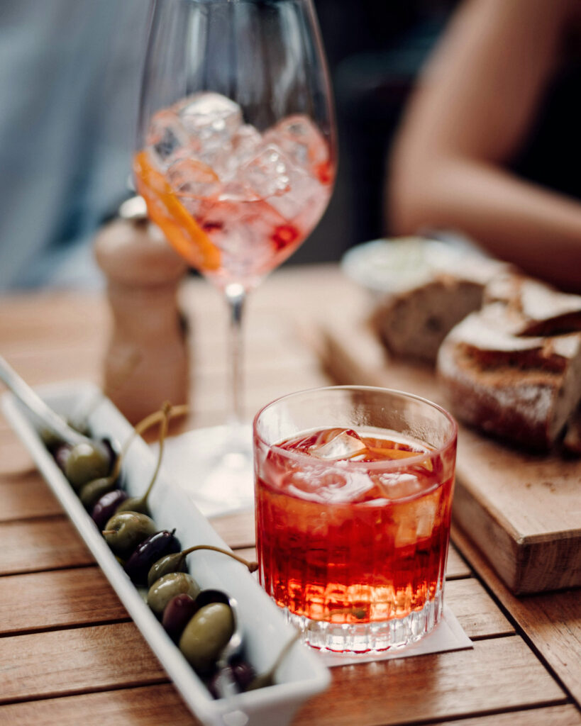 Ein Campari Spritz und ein Glas Aperol Spritz mit Eis auf einem gedeckten Holztisch, flankiert von Oliven und frischem Brot – perfekter Aperitivo-Moment.