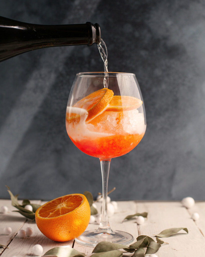 Ein Spritz wird mit Prosecco über Eis und Orangenscheiben aufgegossen – kreatives Food-Styling mit winterlich dekoriertem Hintergrund.