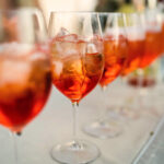 Eine Reihe von Spritzgläsern mit Eiswürfeln auf einer weißen Tischdecke – festlicher Aperitivo-Empfang unter freiem Himmel.