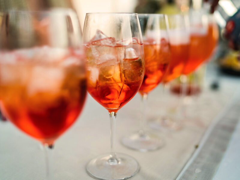 Eine Reihe von Spritzgläsern mit Eiswürfeln auf einer weißen Tischdecke – festlicher Aperitivo-Empfang unter freiem Himmel.
