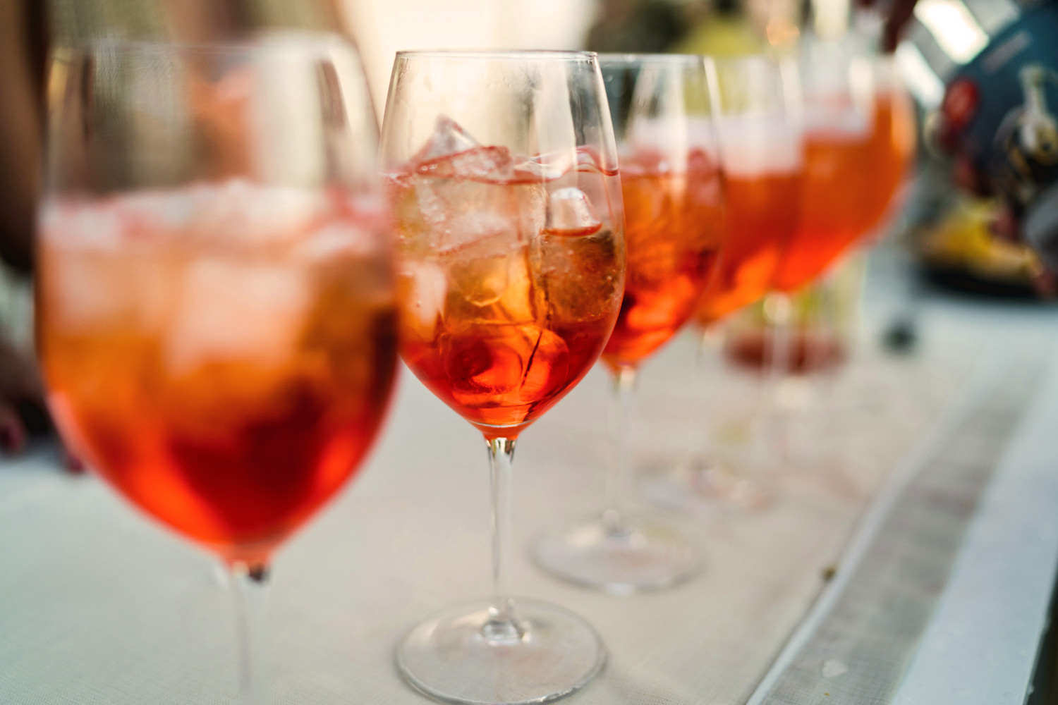Eine Reihe von Spritzgläsern mit Eiswürfeln auf einer weißen Tischdecke – festlicher Aperitivo-Empfang unter freiem Himmel.