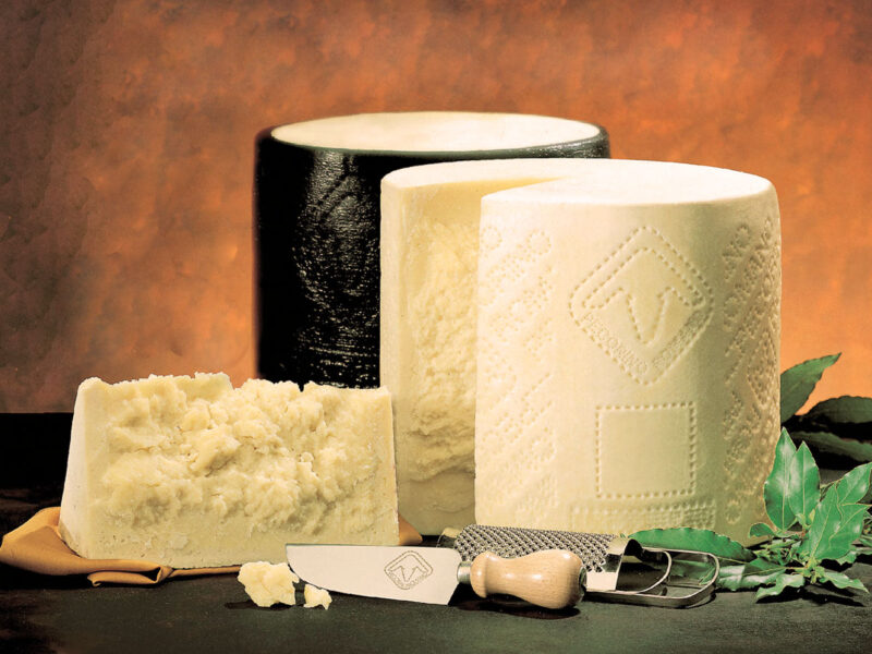 Drei Pecorino-Romano-Laibe in unterschiedlichen Reifegraden – einer mit schwarzer Rinde, zwei aufgebrochen – angerichtet mit Käsemesser, Reibe und Lorbeerblättern auf dunklem Hintergrund; Symbol für die kräftige, salzige Eleganz des römischen Schafskäses.