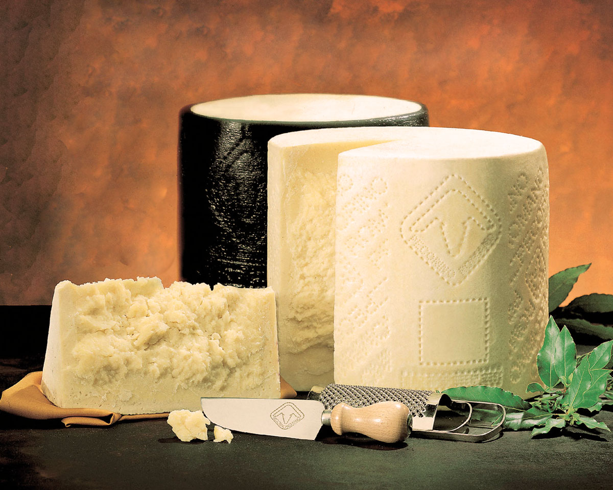 Drei Pecorino-Romano-Laibe in unterschiedlichen Reifegraden – einer mit schwarzer Rinde, zwei aufgebrochen – angerichtet mit Käsemesser, Reibe und Lorbeerblättern auf dunklem Hintergrund; Symbol für die kräftige, salzige Eleganz des römischen Schafskäses.