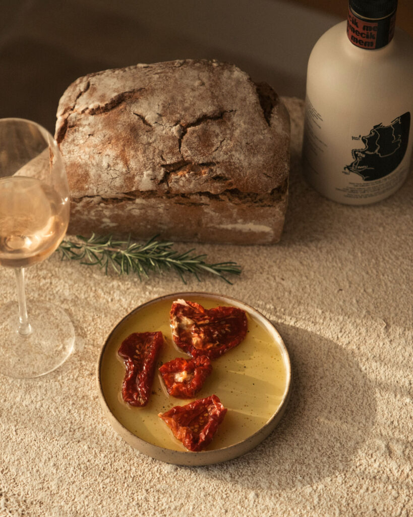 Getrocknete Tomaten in Olivenöl auf einem Keramikteller, serviert mit rustikalem Brot, Rosmarinzweig und einem Glas Roséwein.