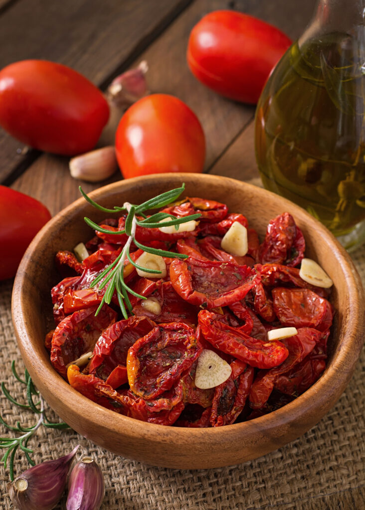 Holzschale mit in Olivenöl eingelegten getrockneten Tomaten, Knoblauchscheiben und frischem Rosmarin – daneben San-Marzano-Tomaten und Knoblauch.