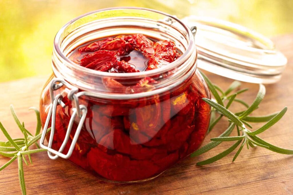 Sonnengereifte, in Öl eingelegte getrocknete Tomaten in einem geöffneten Glas mit Rosmarinzweig – mediterrane Feinkost für Antipasti und Gourmetküche.