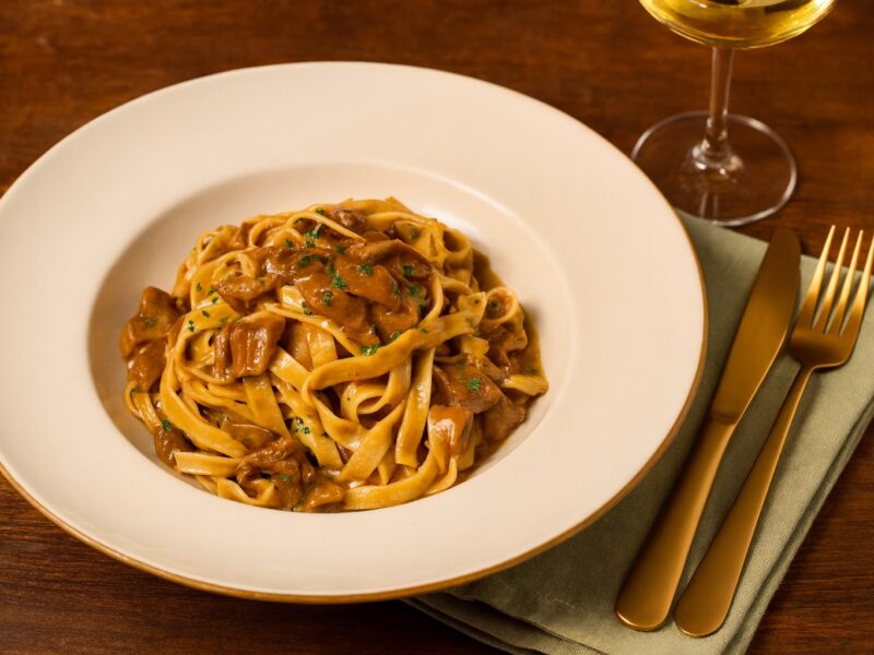 Ein herbstlicher Teller Tagliatelle mit Steinpilzsoße, serviert mit einem Glas Weißwein und goldfarbenem Besteck – Genussmoment pur.