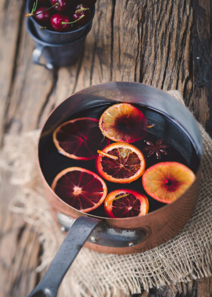 Kupfertopf mit blutroten Orangenscheiben und Vin Brulé auf rustikalem Holztisch mit Sackleinen