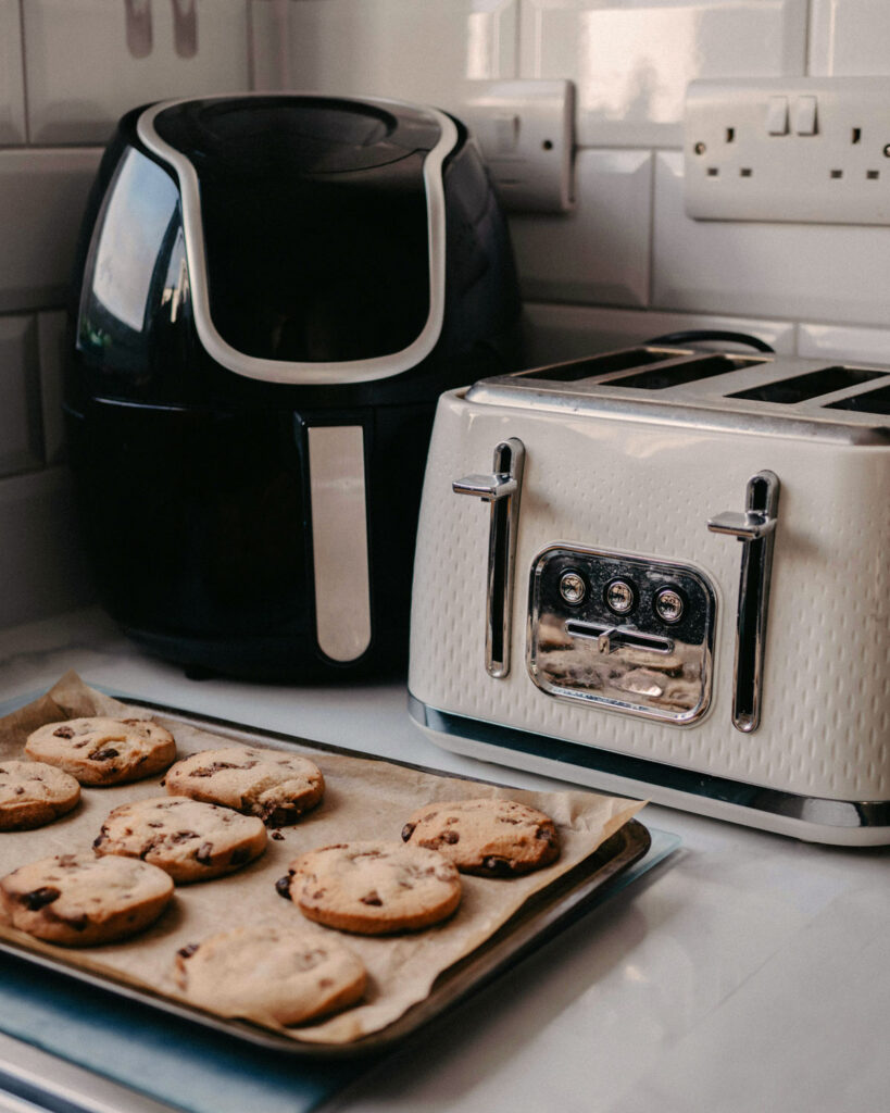 Moderne Heißluftfritteuse in schwarzem Design neben einem Toaster auf einer Küchentheke, im Vordergrund frisch gebackene Cookies auf Backpapier