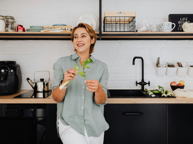 Frau in moderner Küche mit Staudensellerie in der Hand, im Hintergrund eine schwarze Heißluftfritteuse auf der Arbeitsplatte neben einem Induktionskochfeld