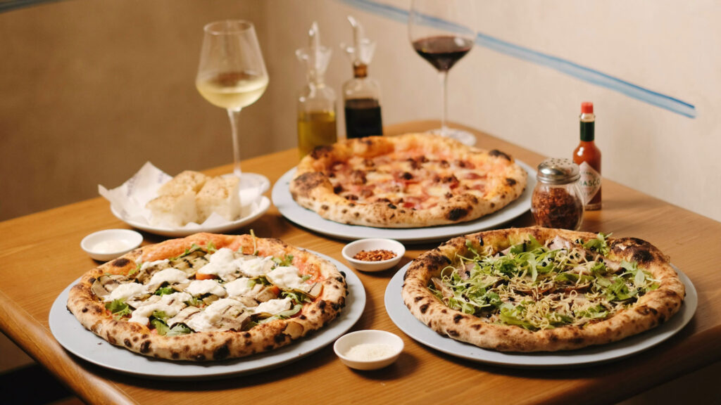 Drei Steinofenpizzen mit verschiedenen Belägen, serviert mit Weißwein, Rotwein und Antipasti – italienische Esskultur in ihrer UNESCO-prämierten Vielfalt