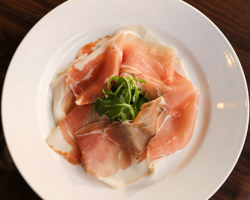 Hauchdünn geschnittener Prosciutto Crudo auf weißem Teller mit Rucola – italienischer Rohschinken als Antipasto.