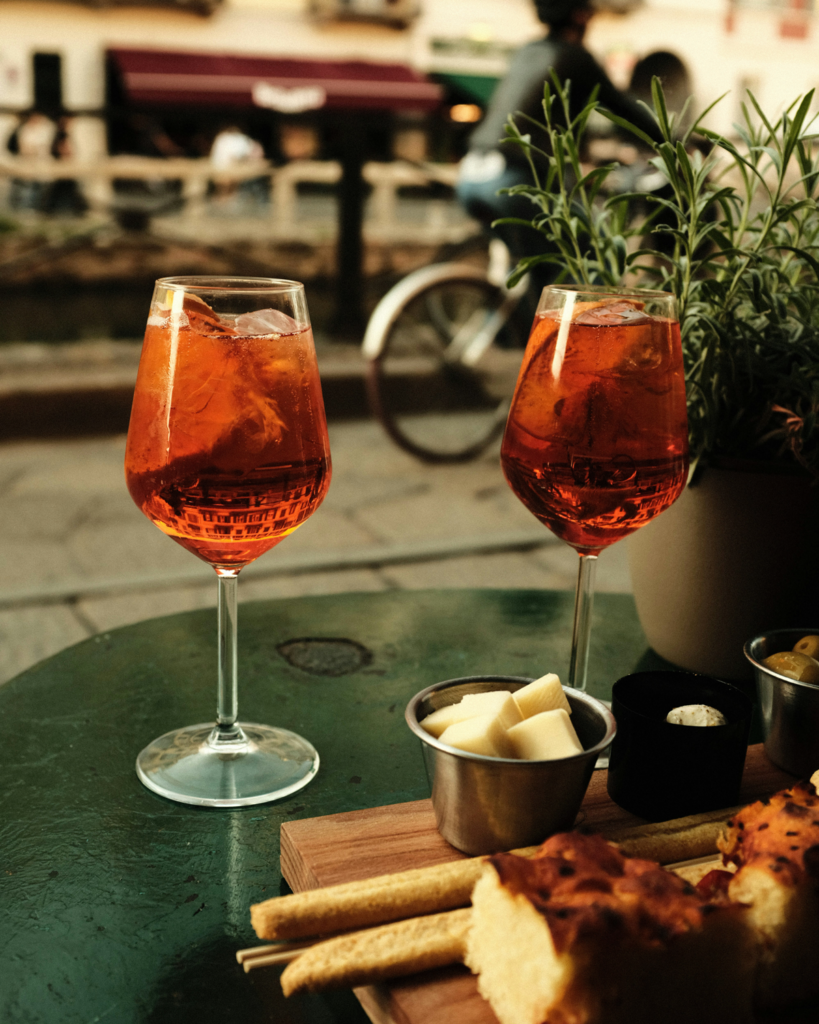 Zwei Gläser Aperol Spritz auf einem Straßencafé-Tisch mit Käse und Snacks – typischer Aperitivo in Italien und Teil der italienischen Aperitivo-Kultur.